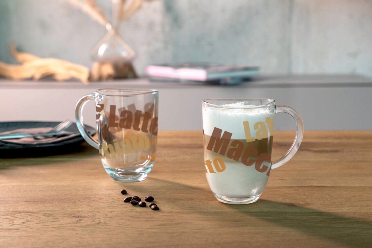Zwei durchsichtige Glastassen mit Henkel, eine gefüllt mit Latte Macchiato, auf einem Holztisch. Im Hintergrund unscharf eine Vase und ein Teller. Perspektive: seitlich.