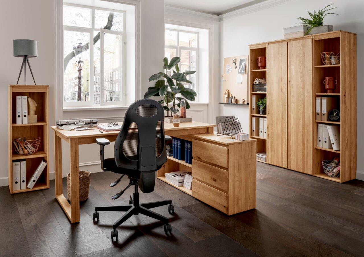 Moderne Büromöbelkombination aus Holz mit Eckschreibtisch, Rollcontainer und Aktenschränken in einem hellen Raum mit großen Fenstern, von vorne betrachtet.