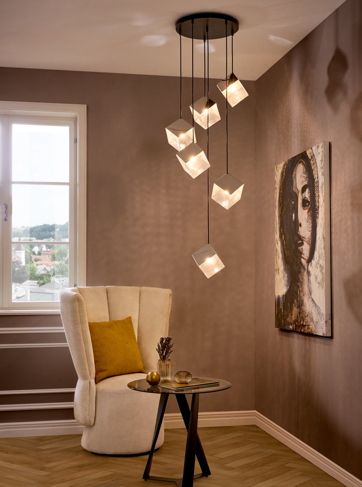 Moderne hanglamp met zes kubusvormige glazen kappen aan een zwarte plafondplaat, die een warm licht verspreidt boven een zithoek met fauteuil en bijzettafel, gefotografeerd vanuit een licht verhoogd perspectief aan de voorkant van de ruimte.