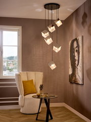 Moderne hanglamp met zes kubusvormige glazen kappen aan een zwarte plafondplaat, die een warm licht verspreidt boven een zithoek met fauteuil en bijzettafel, gefotografeerd vanuit een licht verhoogd perspectief aan de voorkant van de ruimte.