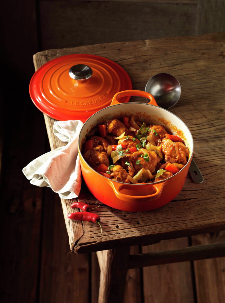 LE CREUSET Bräter Rund 24cm Ofenrot SIGNATURE Runder Bräter in Ofenrot auf einem Holztisch, gefüllt mit einem köstlichen Eintopf. Der Deckel liegt daneben, und die Perspektive ist von oben.