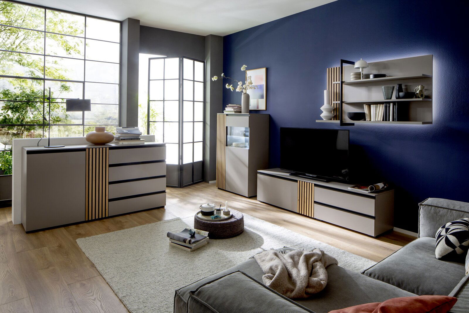 Moderne Wohnzimmermöbel in einem stilvollen Raum mit blauen Wänden, bestehend aus einem Sideboard, einem TV-Lowboard und einem Highboard, mit dekorativen Elementen und einem großen Fenster im Hintergrund. Perspektive: Frontalansicht.