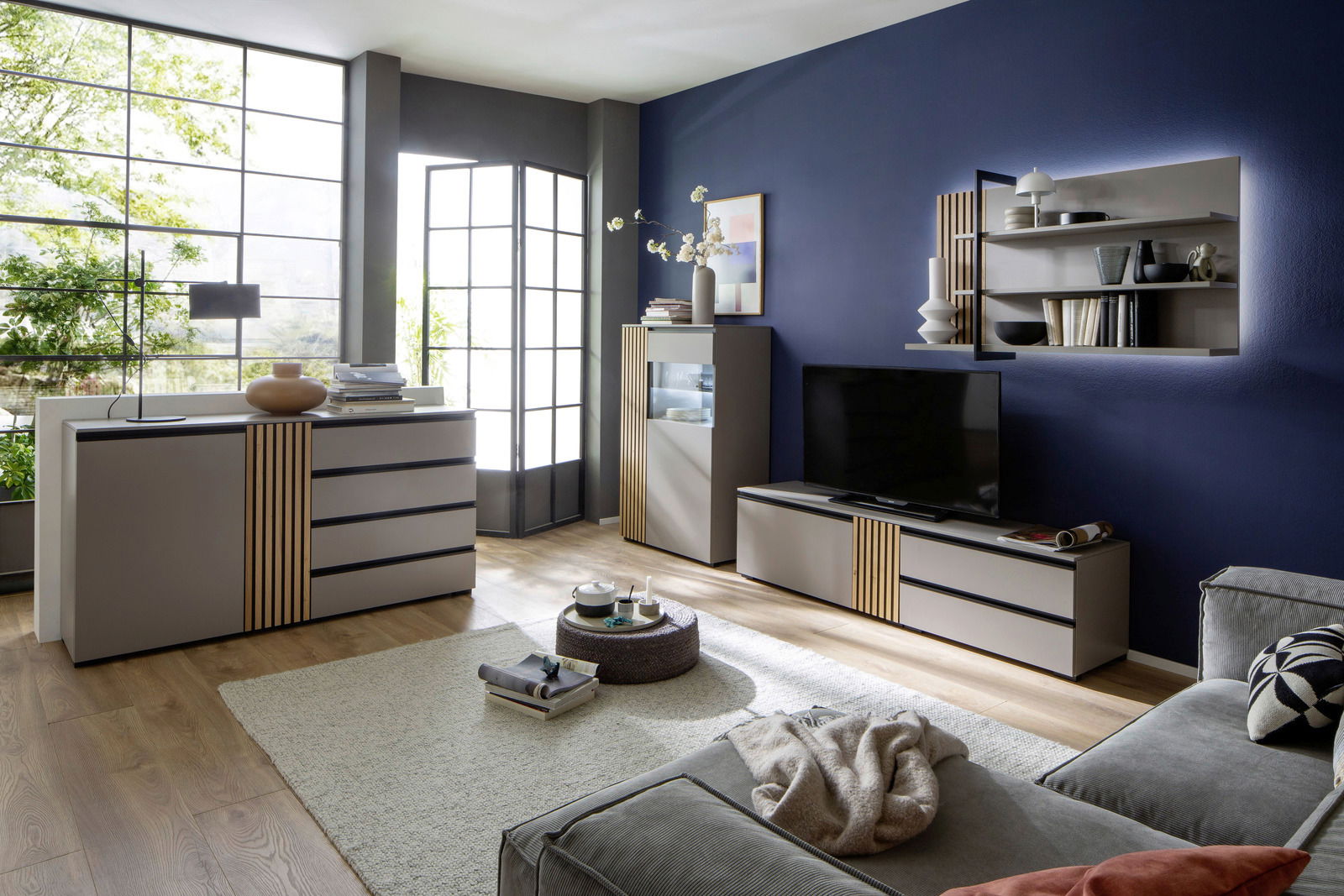 Moderne Wohnzimmermöbel in einem stilvollen Raum mit blauen Wänden, bestehend aus einem Sideboard, einem TV-Lowboard und einem Highboard, mit dekorativen Elementen und einem großen Fenster im Hintergrund. Perspektive: Frontalansicht.