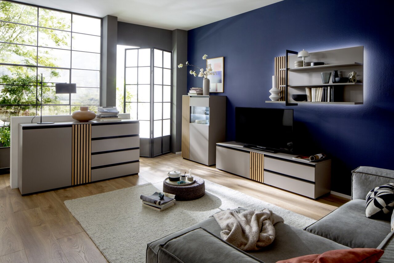 Moderano Tv-kast ARTA Moderne woonkamermeubels in een stijlvolle ruimte met blauwe muren, bestaande uit een dressoir, een tv-kast en een hoge kast, met decoratieve elementen en een groot raam op de achtergrond. Perspectief: vooraanzicht.