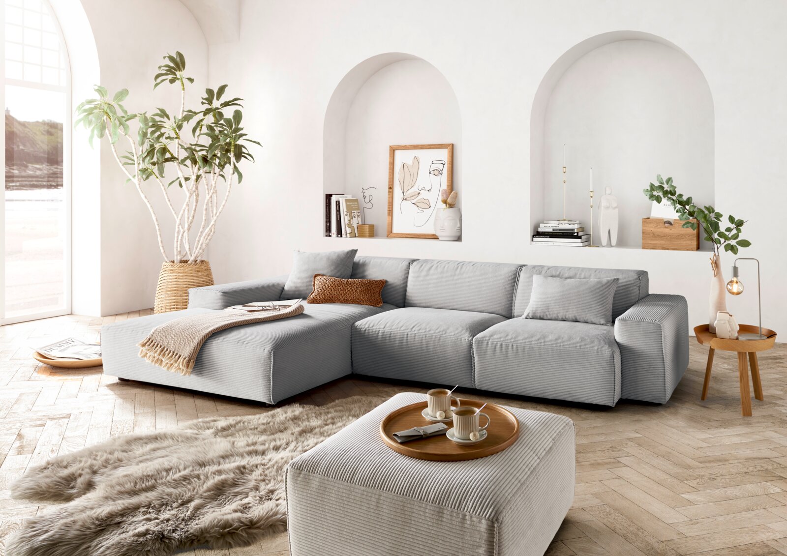 Moderne graue Rundecke in einem hellen Wohnzimmer mit großen Fenstern, Pflanzen und stilvollen Dekorationen, aus der Perspektive von vorne links.