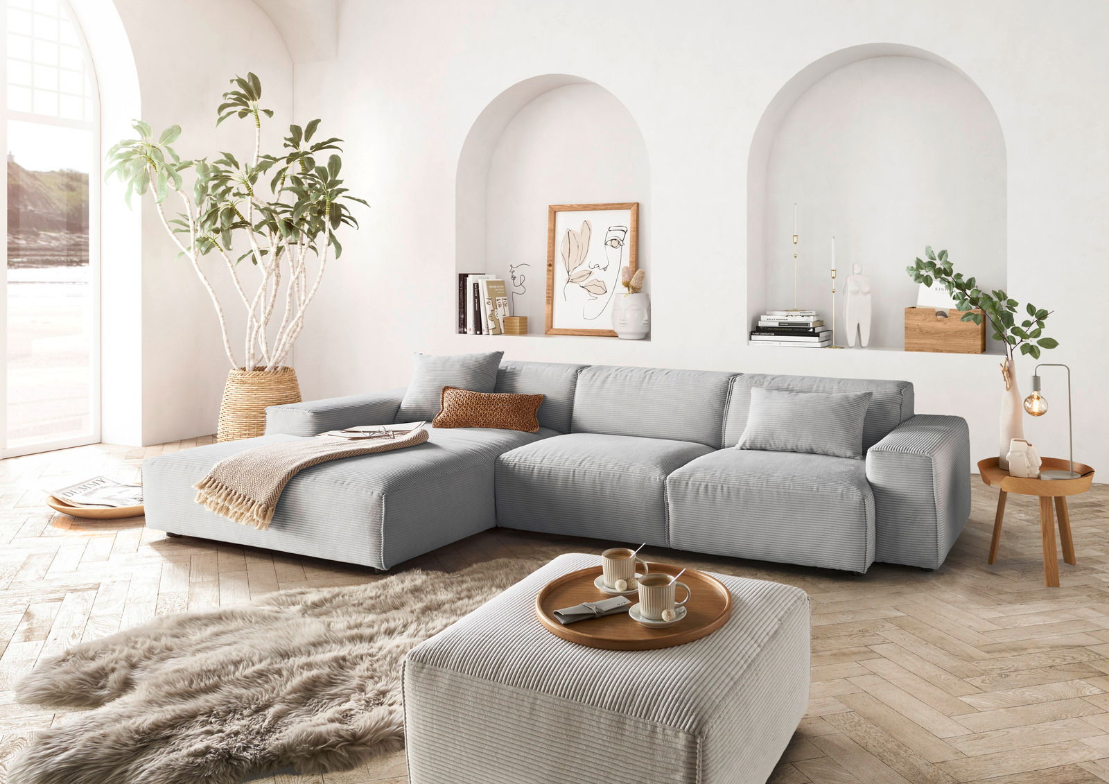 Moderne graue Rundecke in einem hellen Wohnzimmer mit großen Fenstern, Pflanzen und stilvollen Dekorationen, aus der Perspektive von vorne links.