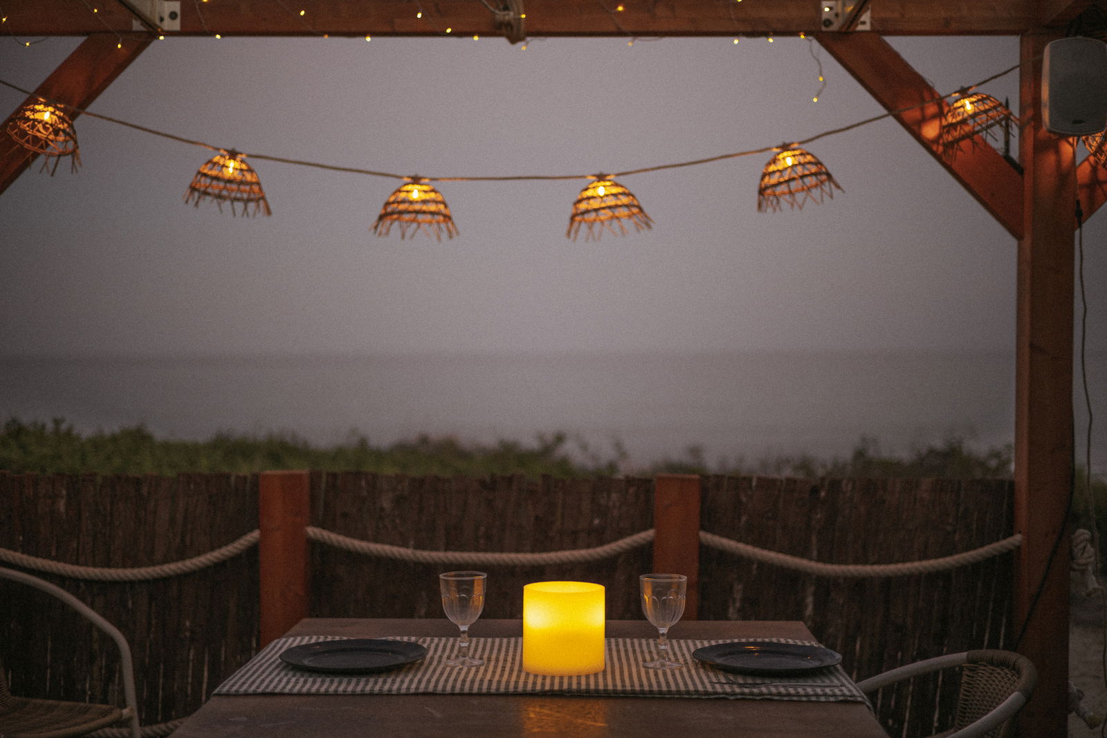 Outdoor-Kerze auf einem gedeckten Tisch mit zwei Gläsern und Tellern, umgeben von stimmungsvoller Beleuchtung in einer abendlichen Strandkulisse, aus der Perspektive eines sitzenden Gastes.