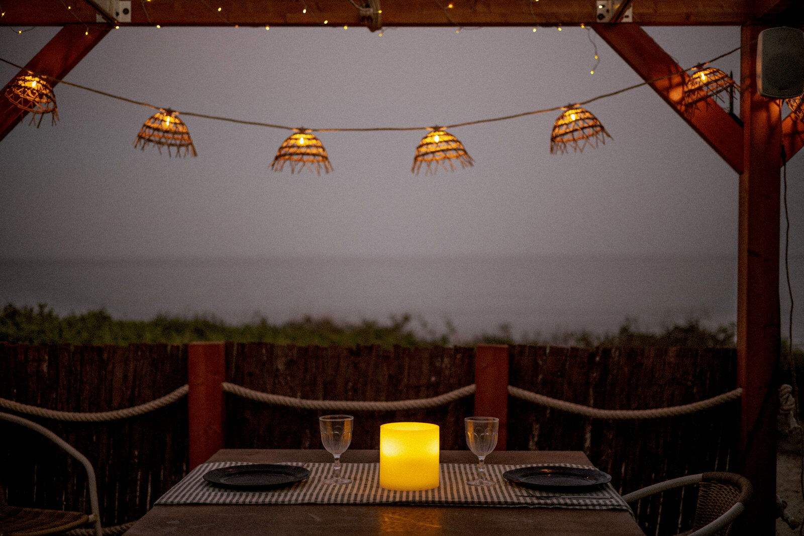 Outdoor-Kerze auf einem gedeckten Tisch mit zwei Gläsern und Tellern, umgeben von stimmungsvoller Beleuchtung in einer abendlichen Strandkulisse, aus der Perspektive eines sitzenden Gastes.