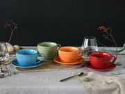 Vier bunte Cappuccinotassen in Blau, Grün, Orange und Rot auf einem Tisch, von vorne fotografiert.
