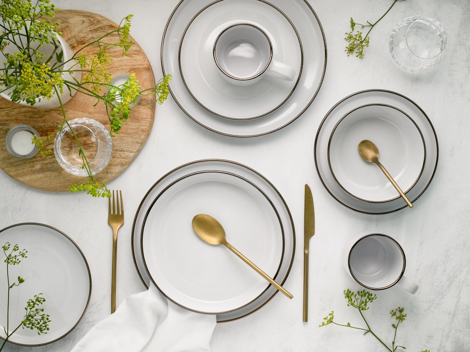 Draufsicht auf ein elegant gedecktes Tischarrangement mit weißem Porzellan-Geschirr mit dunklem Rand, goldfarbenem Besteck, Tassen, Schalen und Tellern auf hellem Untergrund, ergänzt durch Glasgefäße, Holzbrett und grüne Blumen.