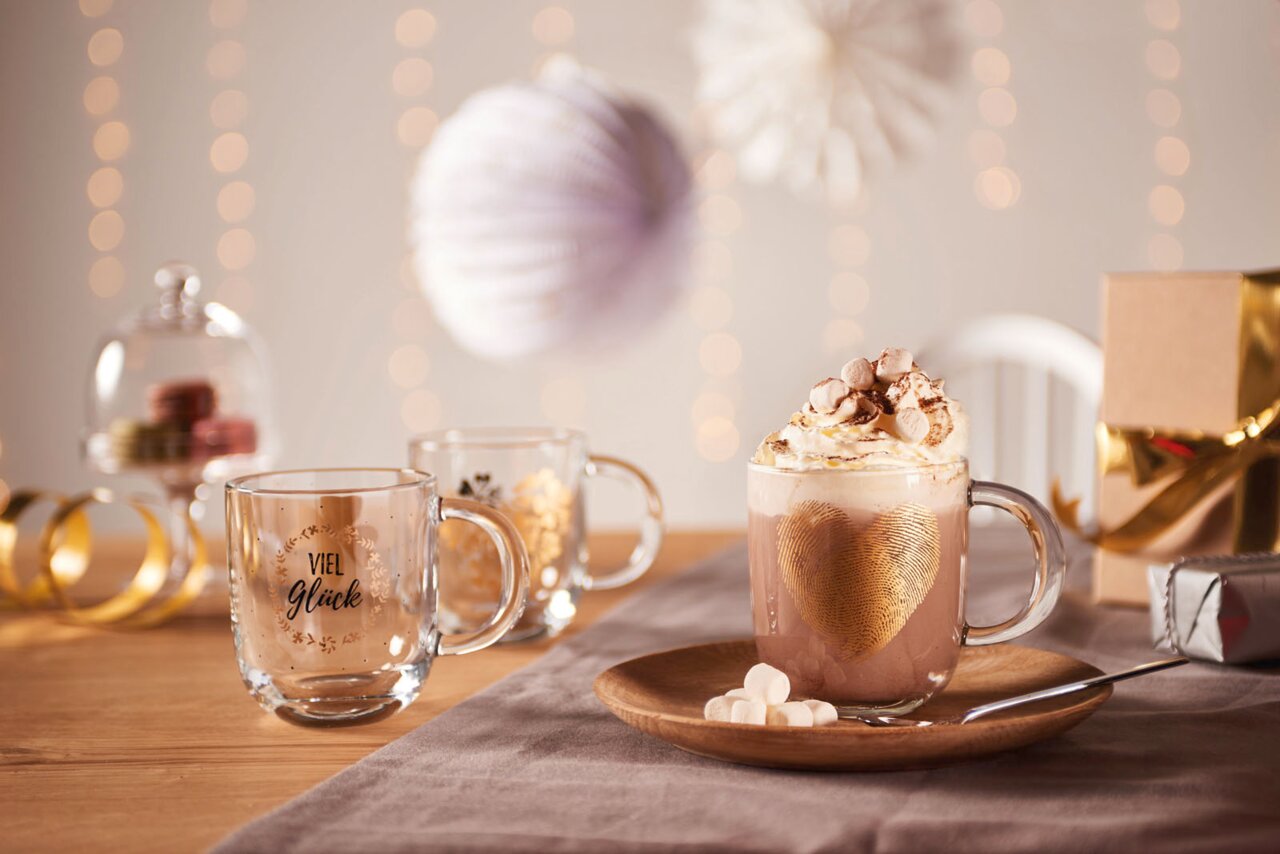 Leonardo Tasse 400ml Herz Drei Glastassen auf einem Tisch, eine mit goldenen Fingerabdruck-Herz und heißer Schokolade, die anderen mit 'Viel Glück' und goldenen Verzierungen, im Hintergrund festliche Dekorationen. Perspektive: seitlich.