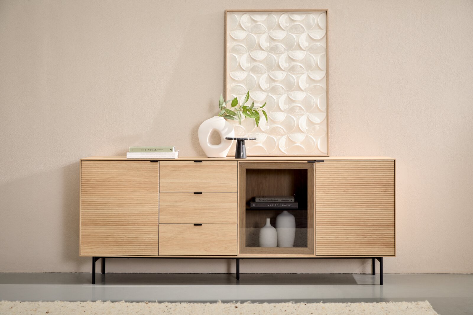 Frontansicht eines modernen Sideboards aus hellem Holz mit schwarzen Metallfüßen, Schubladen, gerillten Türen und einer Vitrine mit Glasfach; dekoriert mit Vase, Büchern und Wandbild im Hintergrund