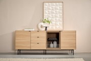 Frontansicht eines modernen Sideboards aus hellem Holz mit schwarzen Metallfüßen, Schubladen, gerillten Türen und einer Vitrine mit Glasfach; dekoriert mit Vase, Büchern und Wandbild im Hintergrund