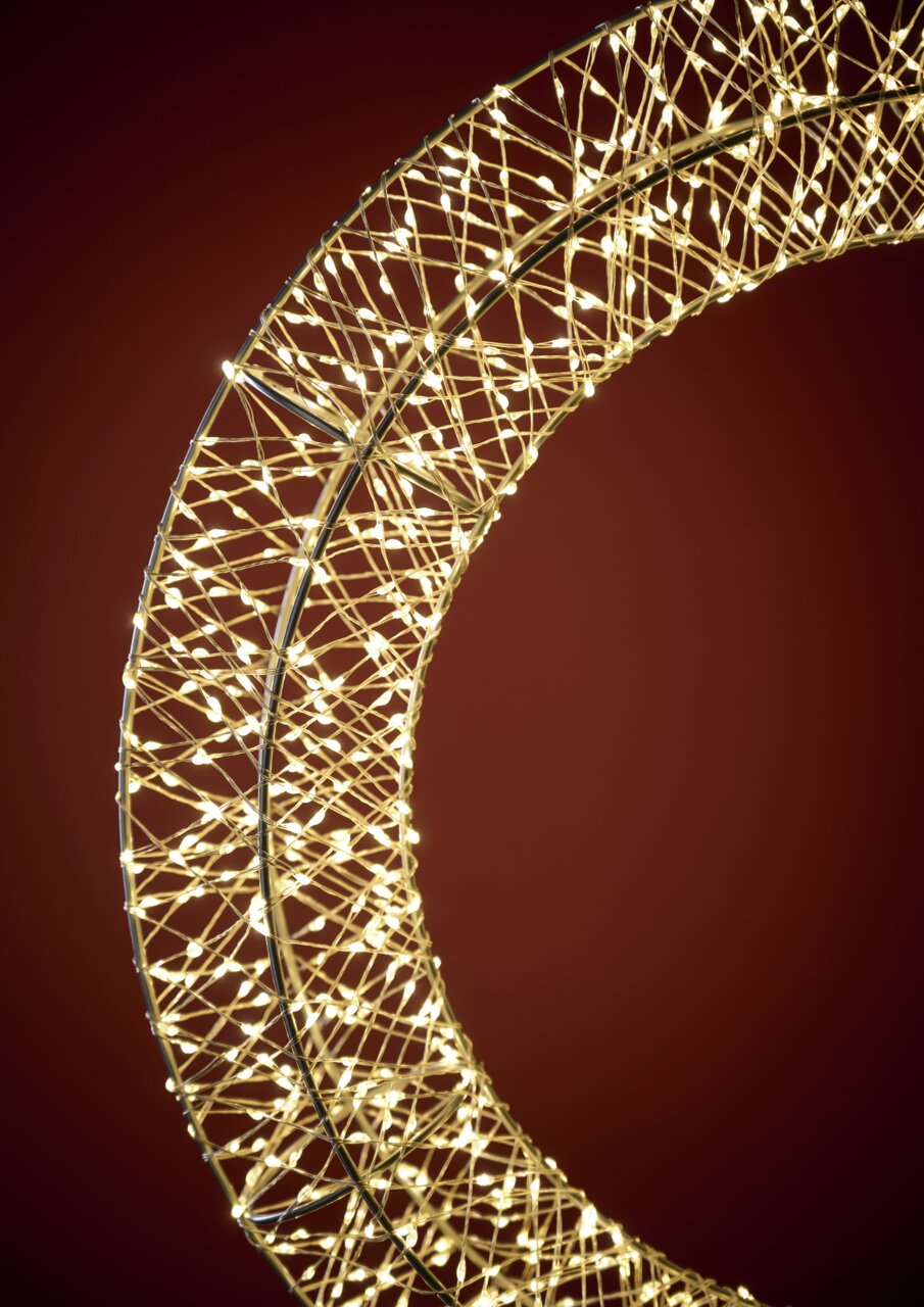 Nahaufnahme eines LED-Kranzes mit einem Durchmesser von 50 cm, der in einem warmen Licht leuchtet. Der Kranz ist aus einem filigranen Drahtgeflecht gefertigt und wird von zahlreichen kleinen LED-Lichtern erhellt. Die Perspektive zeigt den Kranz von der Se