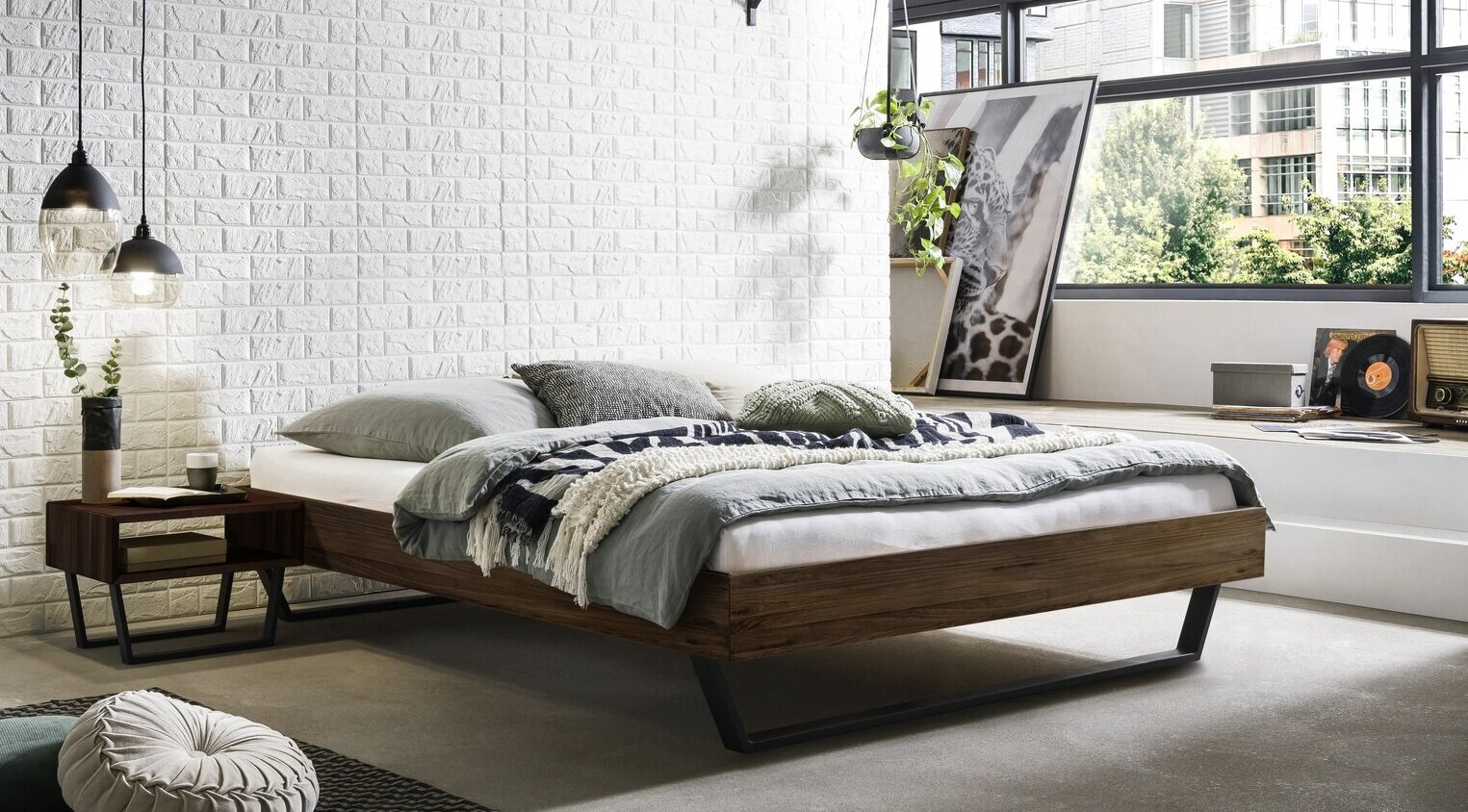 Modern laag houten bed met een zwart metalen onderstel en grijs beddengoed in een lichte loft-slaapkamer met een witte bakstenen muur en grote ramen perspectief: schuin van linksvoor op het bed