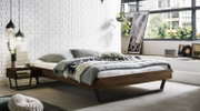 Modern laag houten bed met een zwart metalen onderstel en grijs beddengoed in een lichte loft-slaapkamer met een witte bakstenen muur en grote ramen; perspectief: schuin van linksvoor op het bed