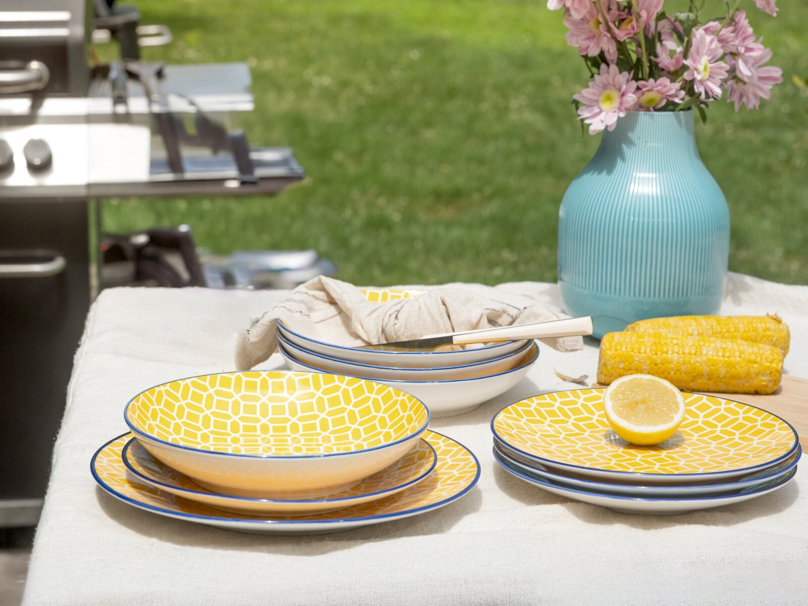 Schräg von vorn aufgenommenes Outdoor-Stillleben mit gelb gemustertem Geschirrset (Teller und Schalen) mit blauem Rand auf weißer Tischdecke, daneben halbierte Zitrone, Maiskolben und blaue Vase mit rosa Blumen im Garten.