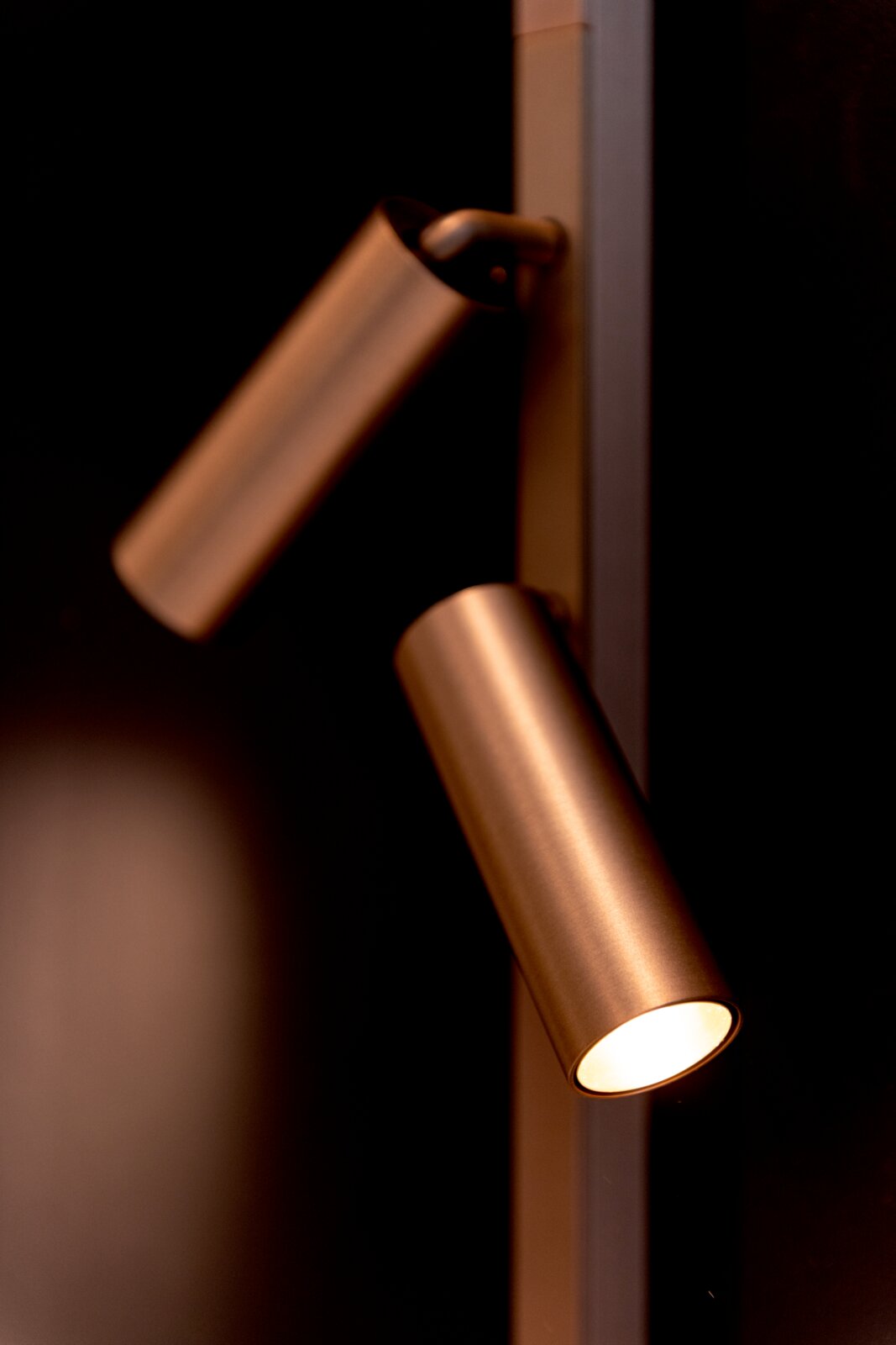 Lucide LED-Schienenstrahler LINIAL Nahaufnahme einer modernen, zylindrischen Wandleuchte/Spotleuchte in Bronze-/Messingoptik mit zwei schwenkbaren Spots, einer leuchtet warmweiß; Perspektive: seitliche Detailansicht schräg von vorn
