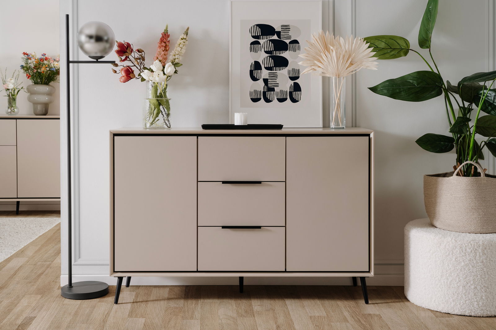 Modernes Sideboard in neutralen Tönen mit drei Schubladen und zwei Türen, dekoriert mit Blumen und Kunstwerk, aus frontaler Perspektive.
