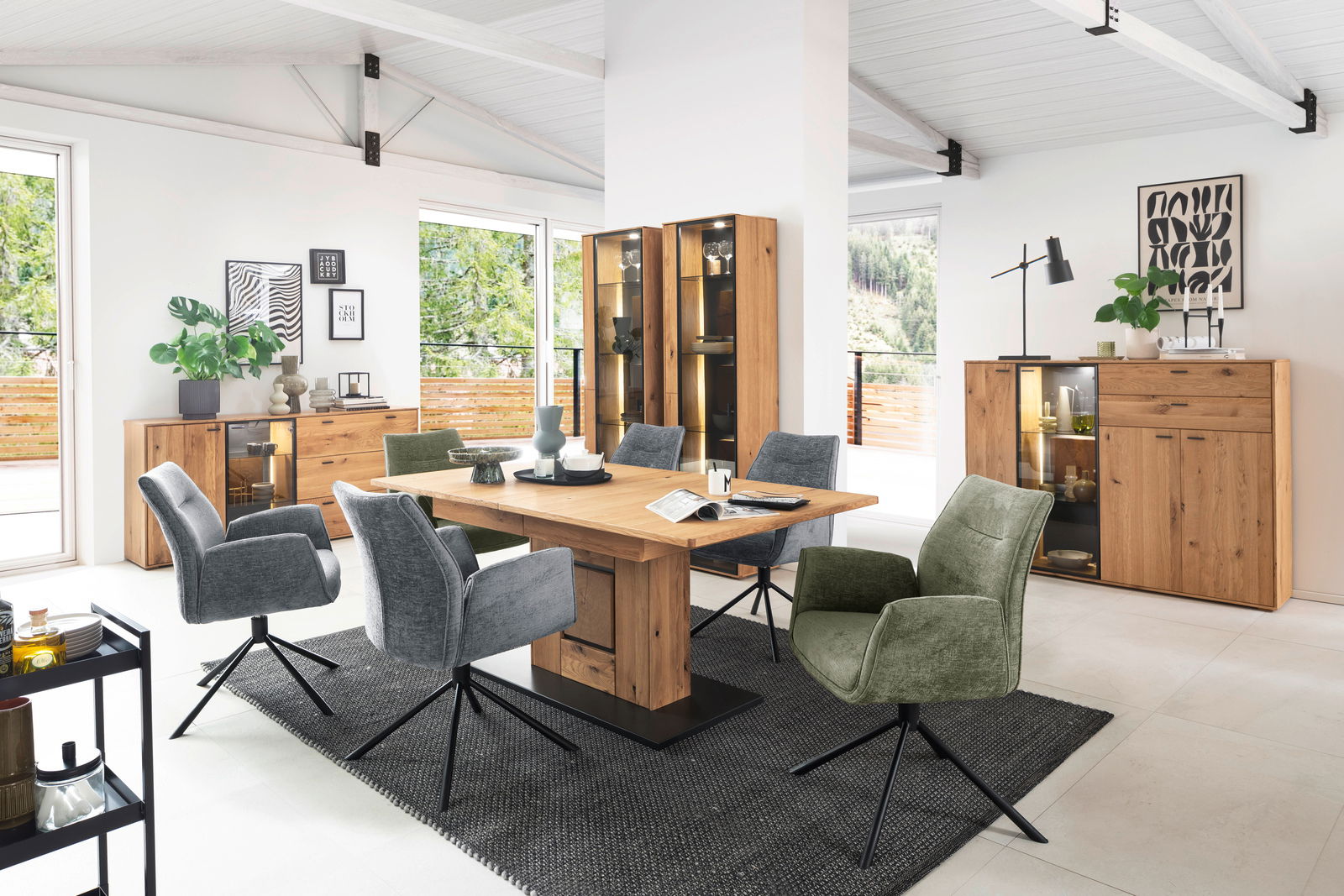 Modernes Esszimmer mit einem massiven Holztisch und Sideboards aus Holz, umgeben von stilvollen, gepolsterten Stühlen in Grau und Grün. Die Perspektive zeigt den Raum von der Seite, mit Blick auf die Fensterfront und die dekorativen Elemente.