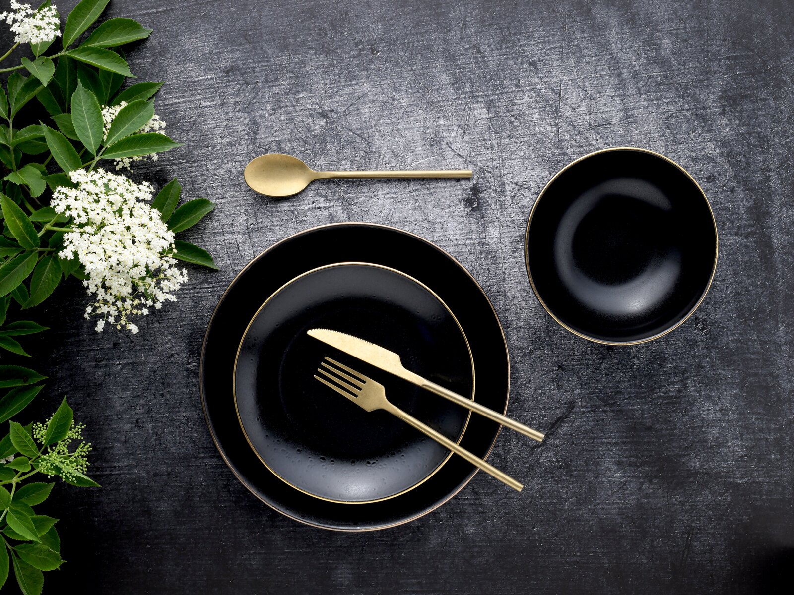 Schwarzes Geschirr-Set mit goldfarbenem Rand (zwei Teller und eine Schale) mit goldfarbenem Besteck auf dunkler Steinoberfläche, dekoriert mit grünen Blättern und weißen Blüten; Ansicht von oben (Flatlay).