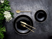 Schwarzes Geschirr-Set mit goldfarbenem Rand (zwei Teller und eine Schale) mit goldfarbenem Besteck auf dunkler Steinoberfläche, dekoriert mit grünen Blättern und weißen Blüten; Ansicht von oben (Flatlay).