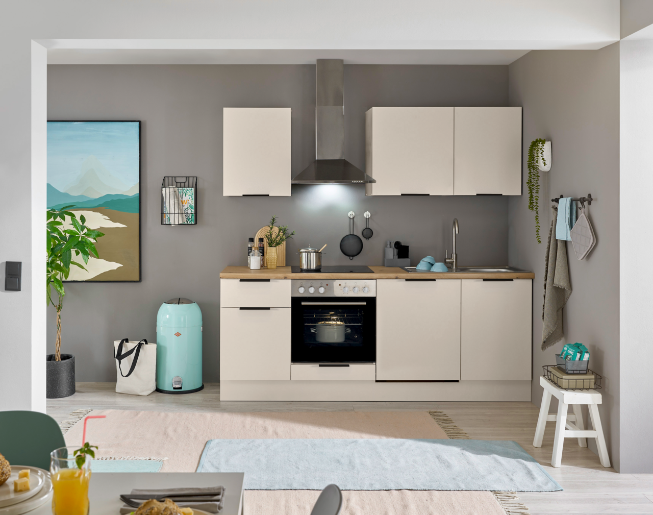 Moderne, kompakte Küchenzeile in Beige mit Holzarbeitsplatte, Backofen, Kochfeld, Dunstabzug und Spüle vor grauer Wand, frontal aus Augenhöhe fotografiert