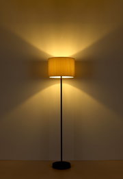 Stehleuchte mit schwarzem Ständer und cremefarbenem Lampenschirm, frontal fotografiert, strahlt warmes Licht an die Wand.