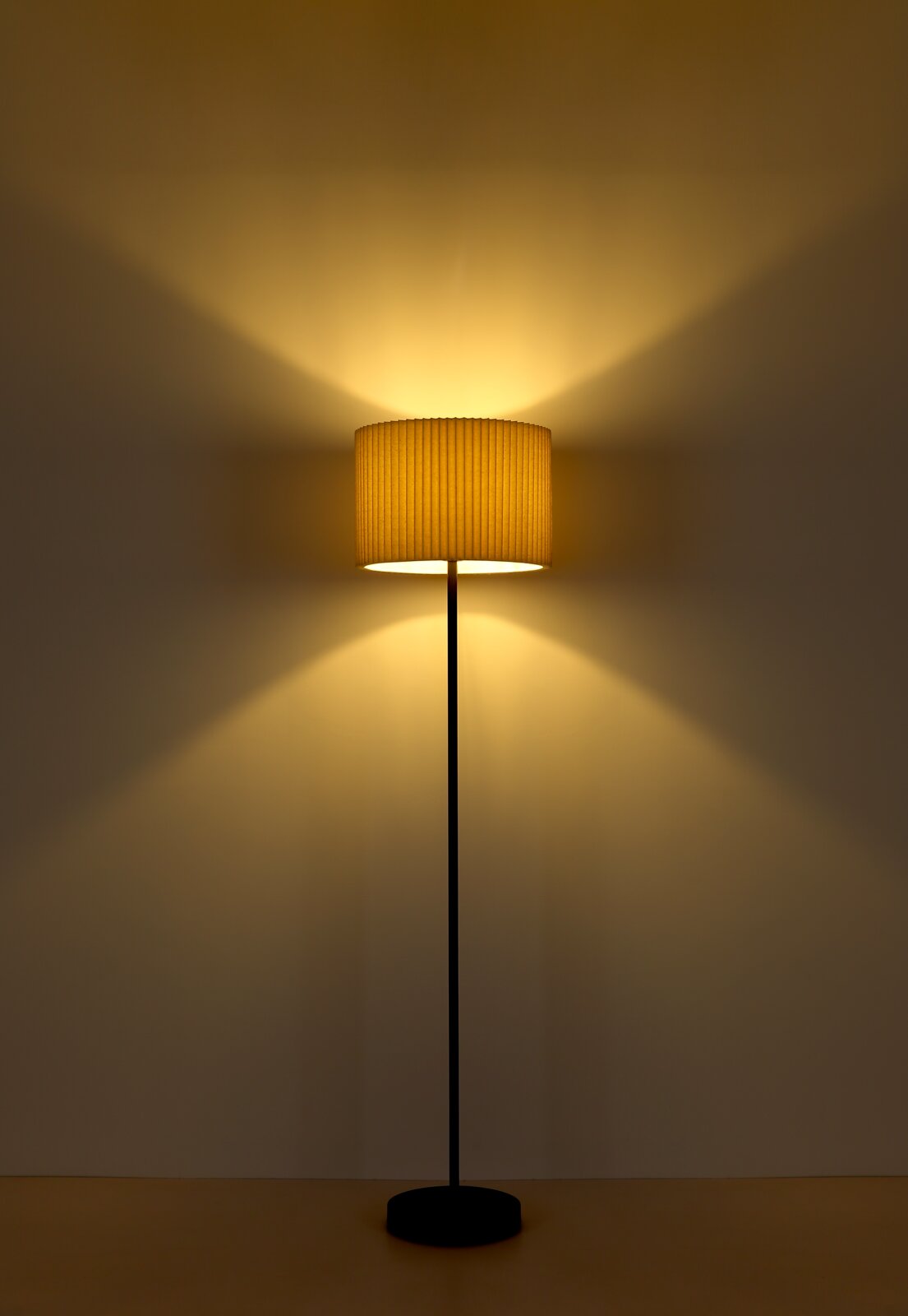 Stehleuchte mit schwarzem Ständer und cremefarbenem Lampenschirm, frontal fotografiert, strahlt warmes Licht an die Wand.