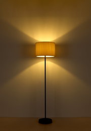 Stehleuchte mit schwarzem Ständer und cremefarbenem Lampenschirm, frontal fotografiert, strahlt warmes Licht an die Wand.
