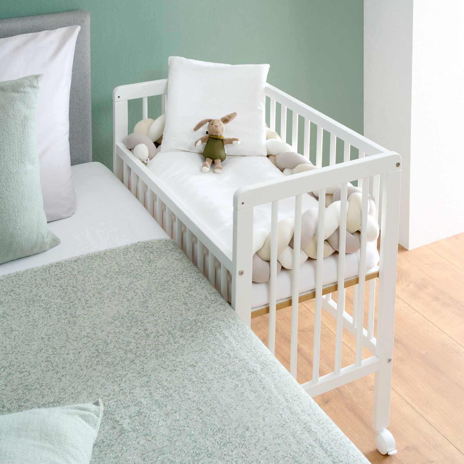 MyBaby Beistellbett NINA Weißes Beistellbett mit Gitterstäben, ausgestattet mit einem Kuscheltier und Kissen, neben einem Bett, aus der Seitenperspektive fotografiert.