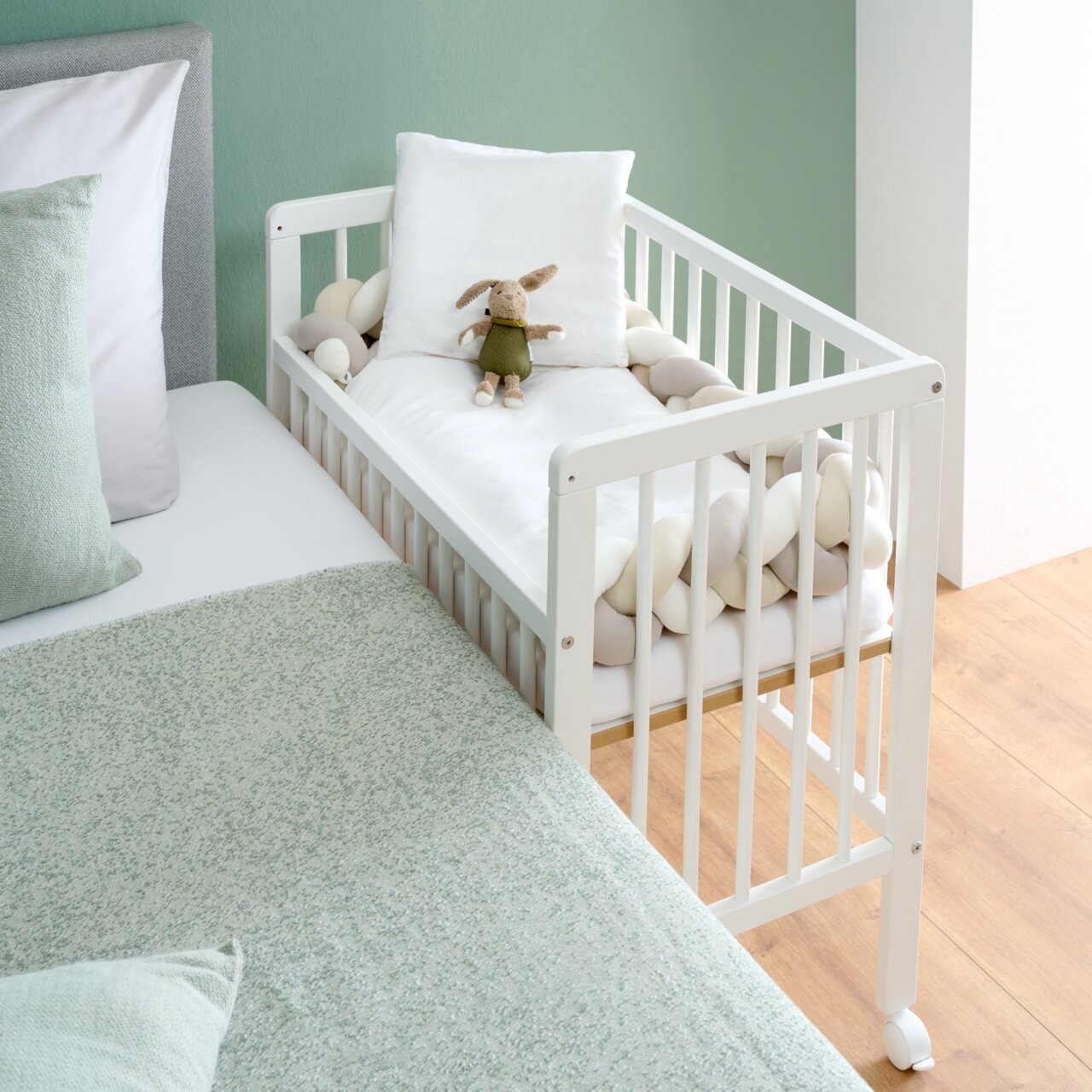 MyBaby Beistellbett NINA Weißes Beistellbett mit Gitterstäben, ausgestattet mit einem Kuscheltier und Kissen, neben einem Bett, aus der Seitenperspektive fotografiert.