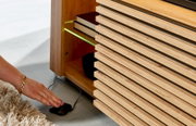 Detailaufnahme aus seitlicher, bodennaher Perspektive: Holz-Sideboard mit Lamellenfront und offenem Fach, grün beleuchtete Glasablage mit Büchern; Hand bedient Fußschalter am Boden