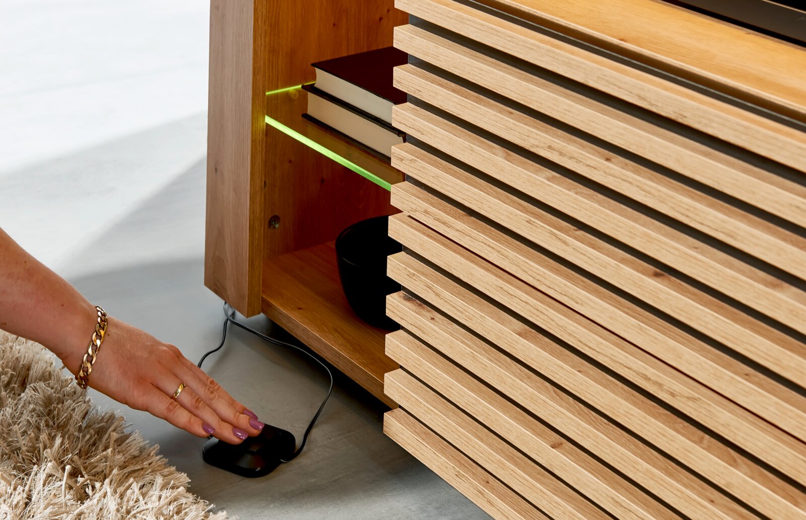 Detailaufnahme aus seitlicher, bodennaher Perspektive: Holz-Sideboard mit Lamellenfront und offenem Fach, grün beleuchtete Glasablage mit Büchern; Hand bedient Fußschalter am Boden