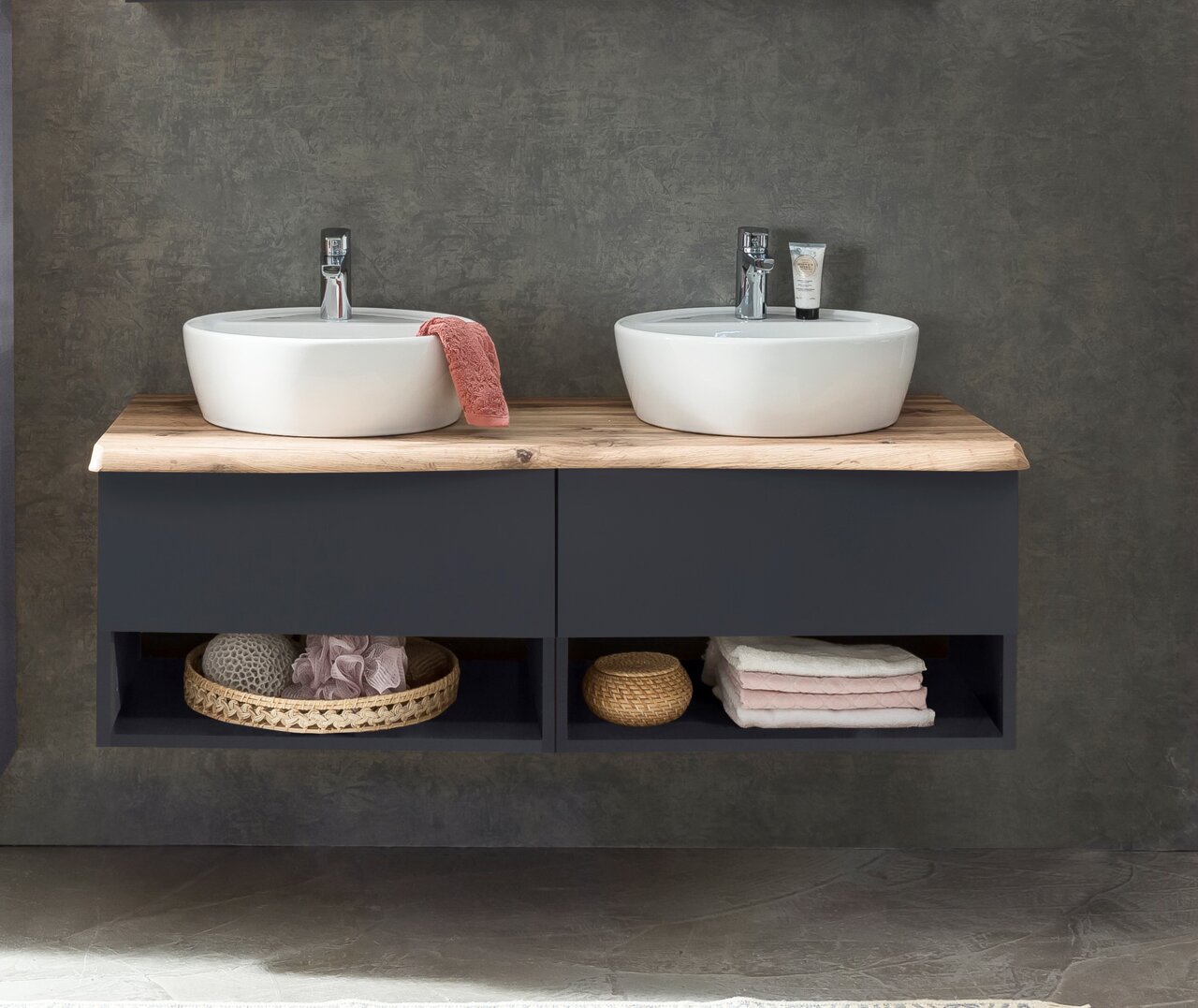 Frontale Ansicht eines modernen Waschtisch-Sets mit zwei runden Waschbecken auf einer Holzplatte, darunter dunkle Schubladen und offene Fächer mit Handtüchern und Körben.
