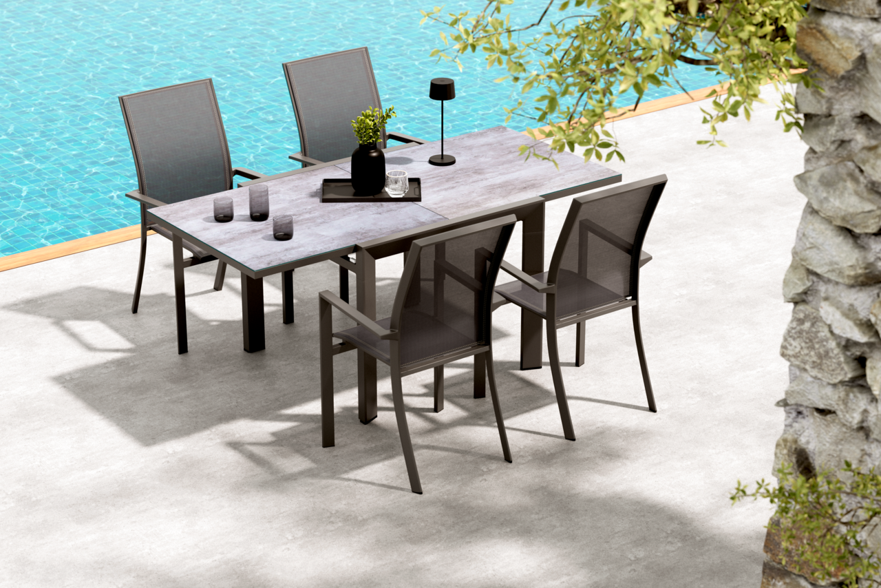 Moderner Outdoor-Esstisch mit grauer Steinoptik-Tischplatte und dunklem Gestell, umgeben von vier passenden Gartenstühlen mit Textilbespannung, auf einer Terrasse am Pool; Perspektive: leicht erhöhte Schrägansicht von vorne.
