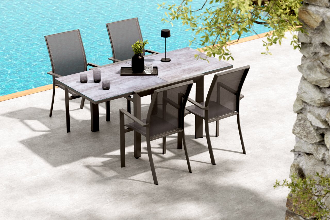 Moderner Outdoor-Esstisch mit grauer Steinoptik-Tischplatte und dunklem Gestell, umgeben von vier passenden Gartenstühlen mit Textilbespannung, auf einer Terrasse am Pool; Perspektive: leicht erhöhte Schrägansicht von vorne.