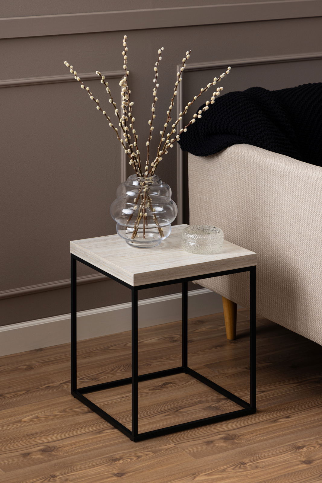 Moderner Couchtisch mit schwarzem Metallgestell und heller Holzplatte, seitliche Perspektive, dekoriert mit einer Glasvase und einem kleinen Glasobjekt.