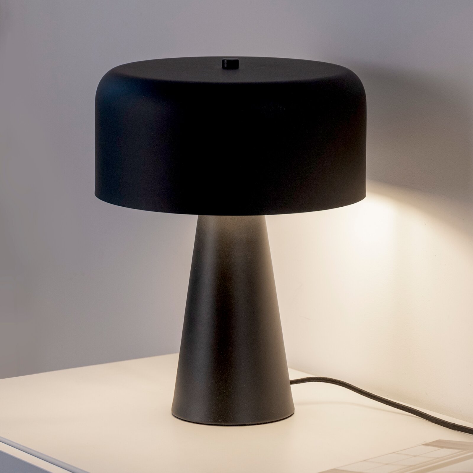 Schwarze, moderne Tischlampe mit breitem, rundem Schirm und konischem Fuß auf hellem Tisch vor grauer Wand, frontal in leichter Seitenansicht fotografiert