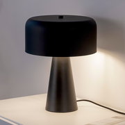 Schwarze, moderne Tischlampe mit breitem, rundem Schirm und konischem Fuß auf hellem Tisch vor grauer Wand, frontal in leichter Seitenansicht fotografiert