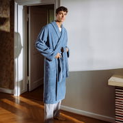 Mann in einem blauen Bademantel mit Schalkragen, stehend in einem Raum mit Holzboden, von vorne betrachtet.