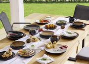 Ein stilvoll gedeckter Tisch im Freien mit einem 16-teiligen Kombiservice in dunklen Tönen, bestehend aus Tellern, Schalen und Tassen, dekoriert mit frischen Speisen und Kräutern, aus der Vogelperspektive fotografiert.