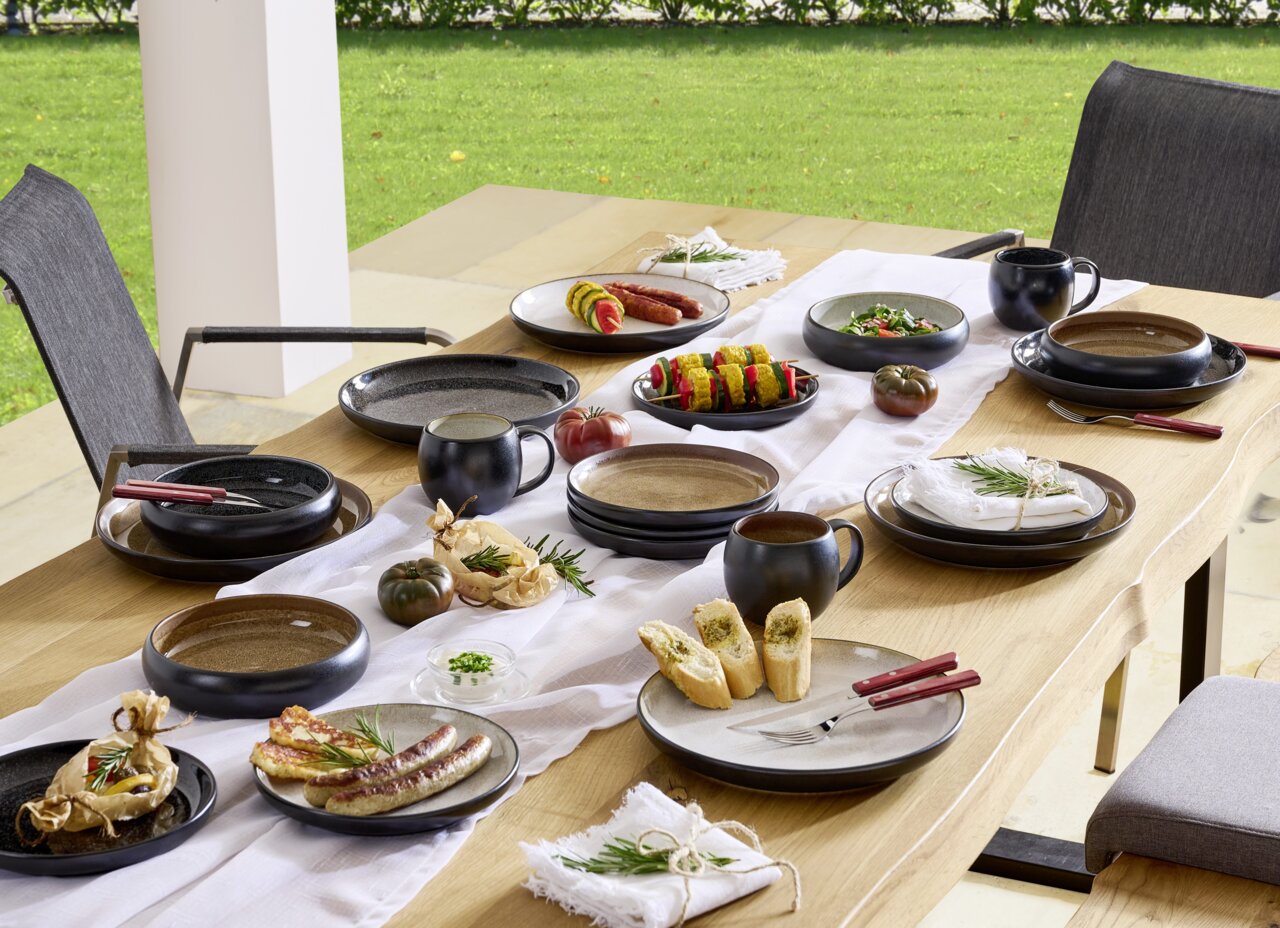 Ein stilvoll gedeckter Tisch im Freien mit einem 16-teiligen Kombiservice in dunklen Tönen, bestehend aus Tellern, Schalen und Tassen, dekoriert mit frischen Speisen und Kräutern, aus der Vogelperspektive fotografiert.