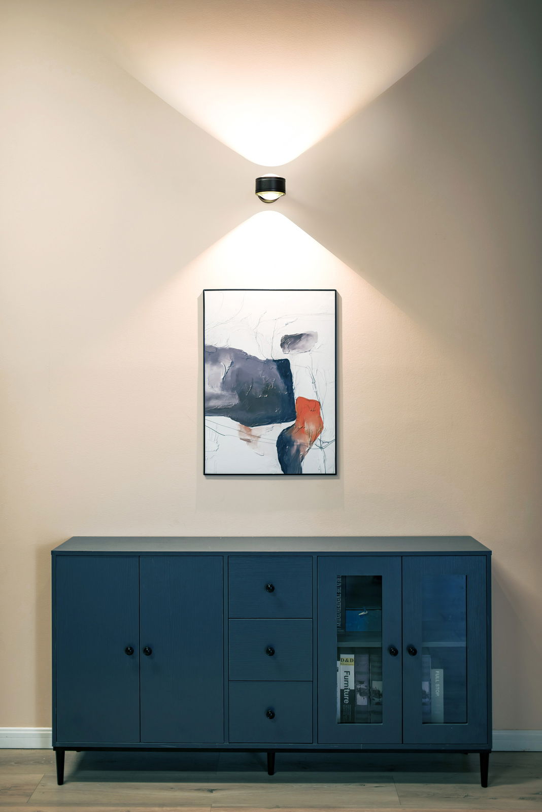 Moderne LED Wandleuchte in einem stilvollen Raum, frontal betrachtet, über einem Kunstwerk und einer blauen Kommode montiert.