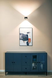 Moderne LED Wandleuchte in einem stilvollen Raum, frontal betrachtet, über einem Kunstwerk und einer blauen Kommode montiert.