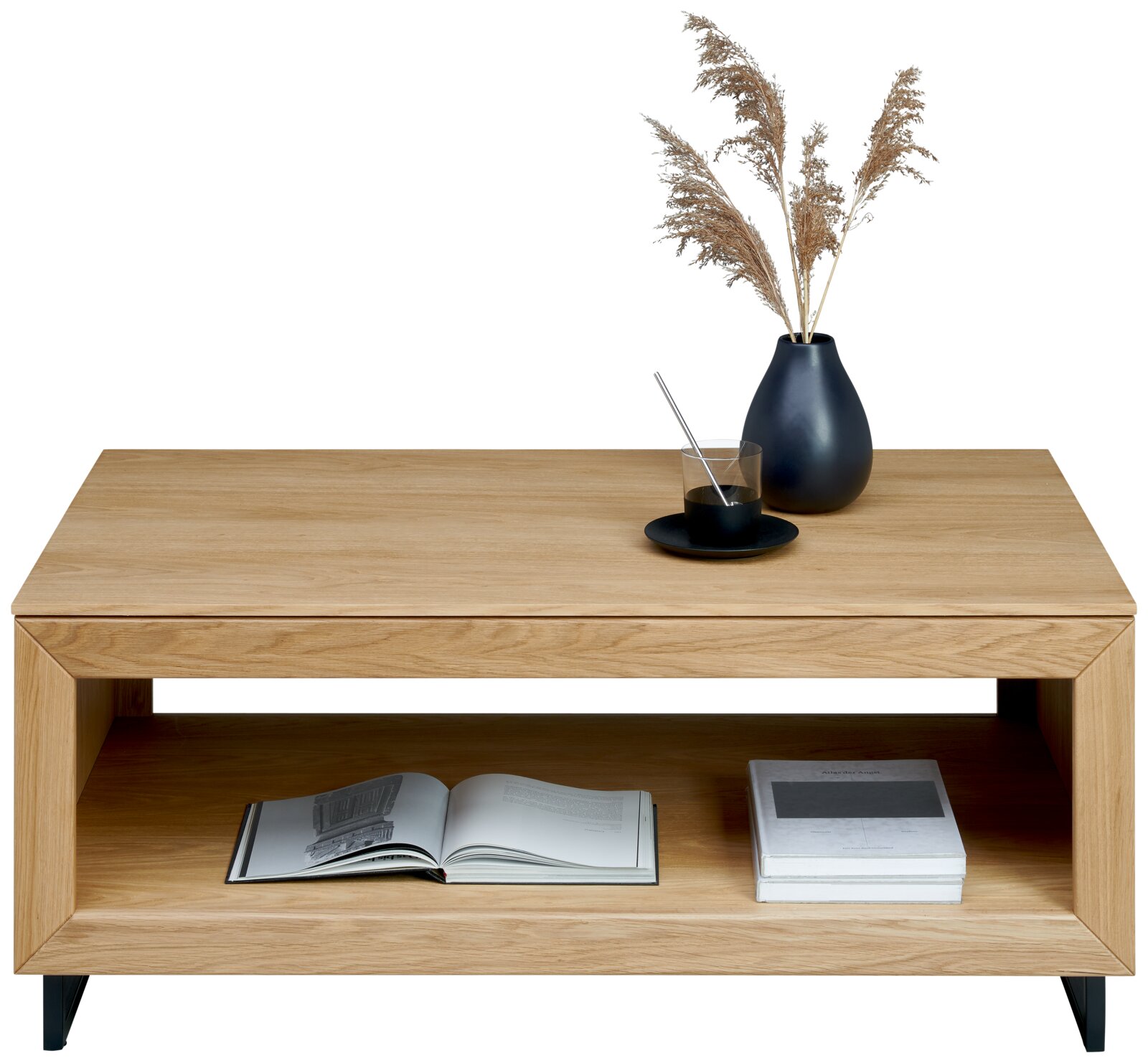Licht eikenhouten salontafel met open vak en zwarte metalen poten, gefotografeerd in frontaal aanzicht; op het blad een zwarte vaas met droogpluimen en een glas op schoteltje, in het vak een open boek en stapel boeken