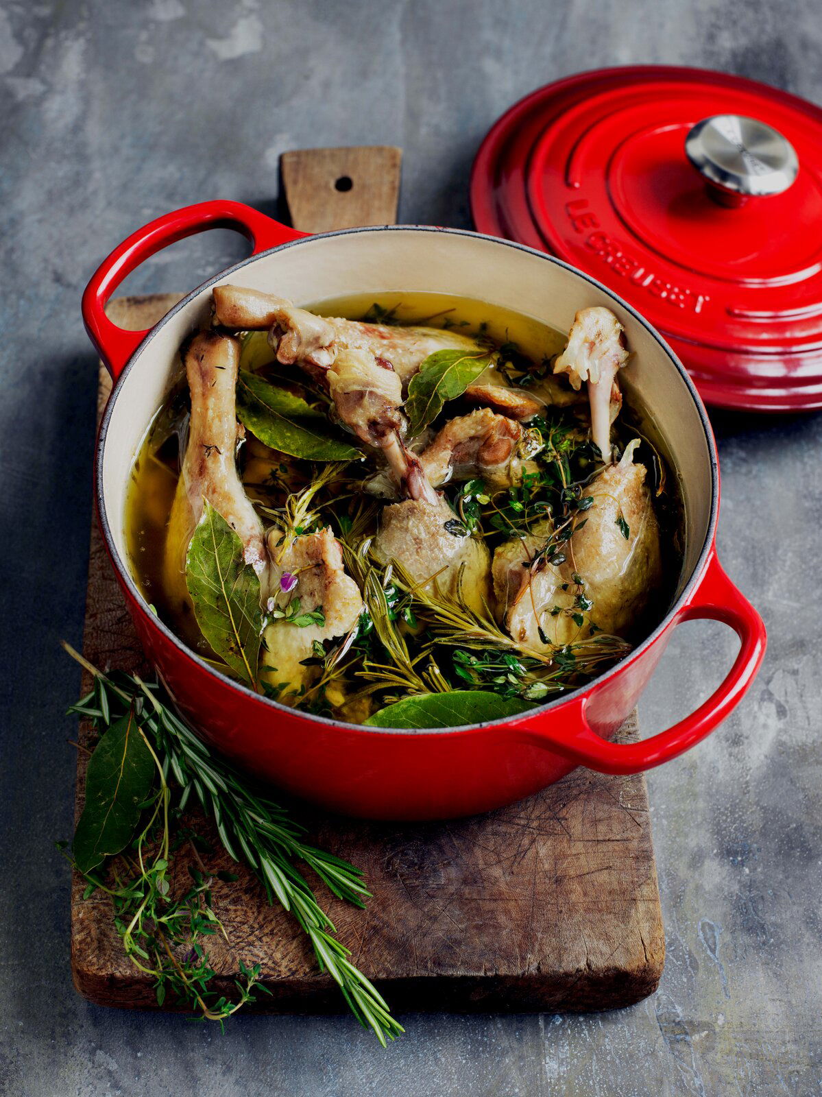 Roter Le Creuset Bräter, gefüllt mit Fleisch und Kräutern, auf einem Holzbrett von oben fotografiert.