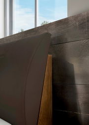 Nahaufnahme eines dunkelbraunen, gepolsterten Bettkopfteils mit Holzrahmen vor dunkler Holzpaneelwand, seitliche Perspektive von links