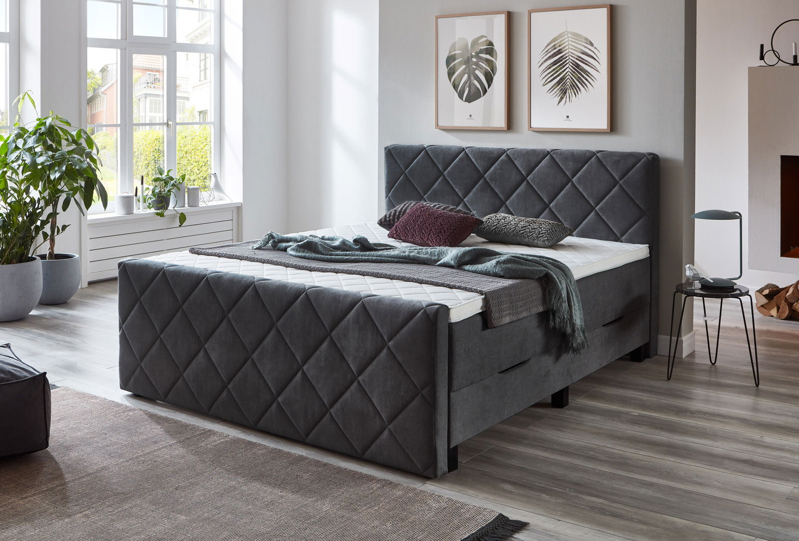 Graues Boxspringbett mit gestepptem Kopf- und Fußteil in einem modernen Schlafzimmer, seitliche Perspektive.
