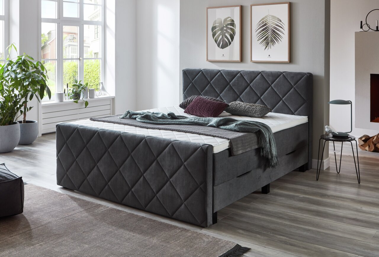 Grijs boxspringbed met doorgestikt hoofd- en voeteneinde in een moderne slaapkamer, gezien vanaf de zijkant.